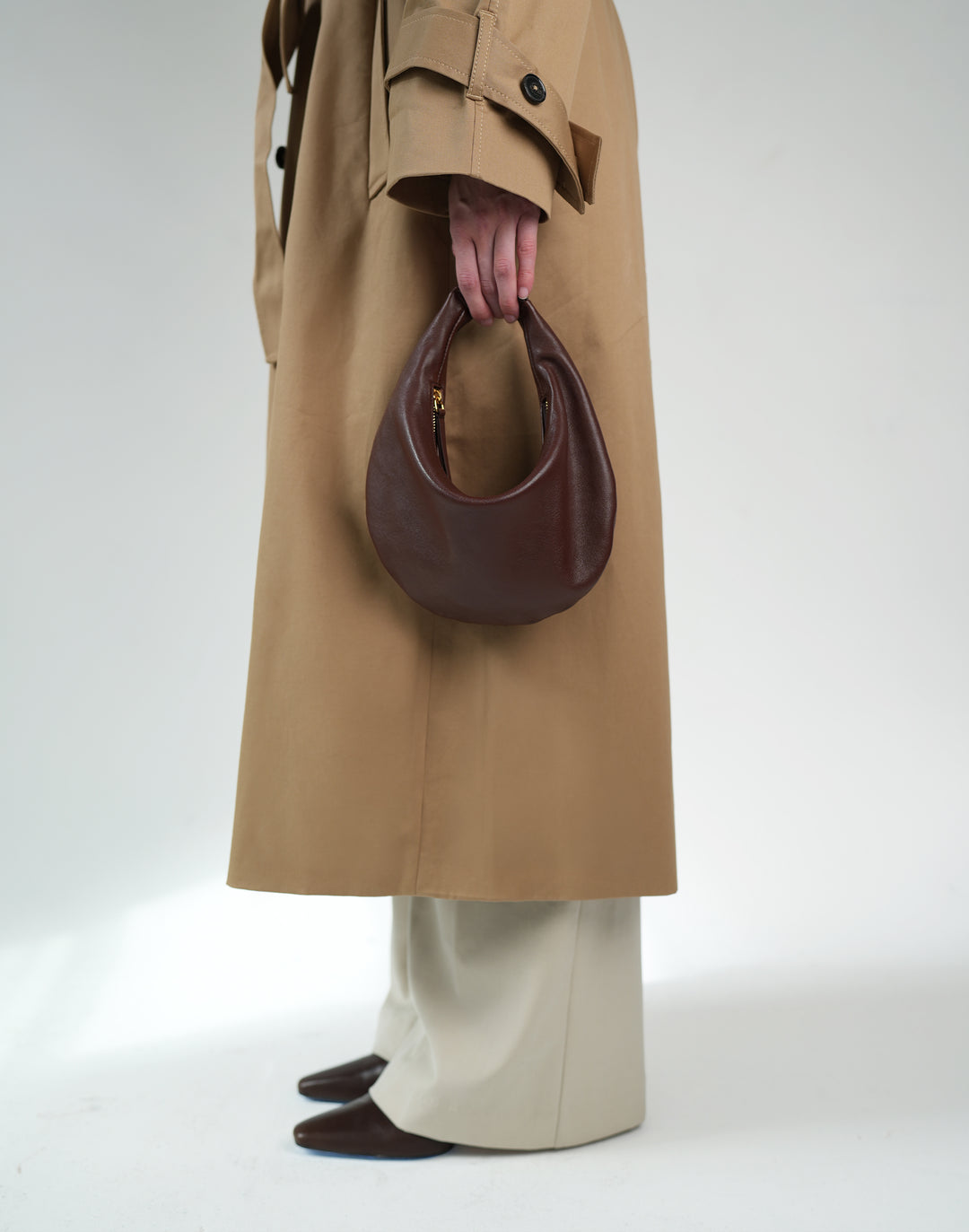 A person wearing a long tan trench coat, cream wide-leg trousers, and dark brown pointed shoes holds a dark brown crescent-shaped leather handbag against a plain light gray studio background. #color_bordeaux-vintage