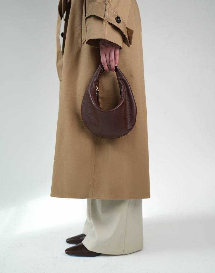 A person wearing a long tan trench coat, cream wide-leg trousers, and dark brown pointed shoes holds a dark brown crescent-shaped leather handbag against a plain light gray studio background. #color_bordeaux-vintage