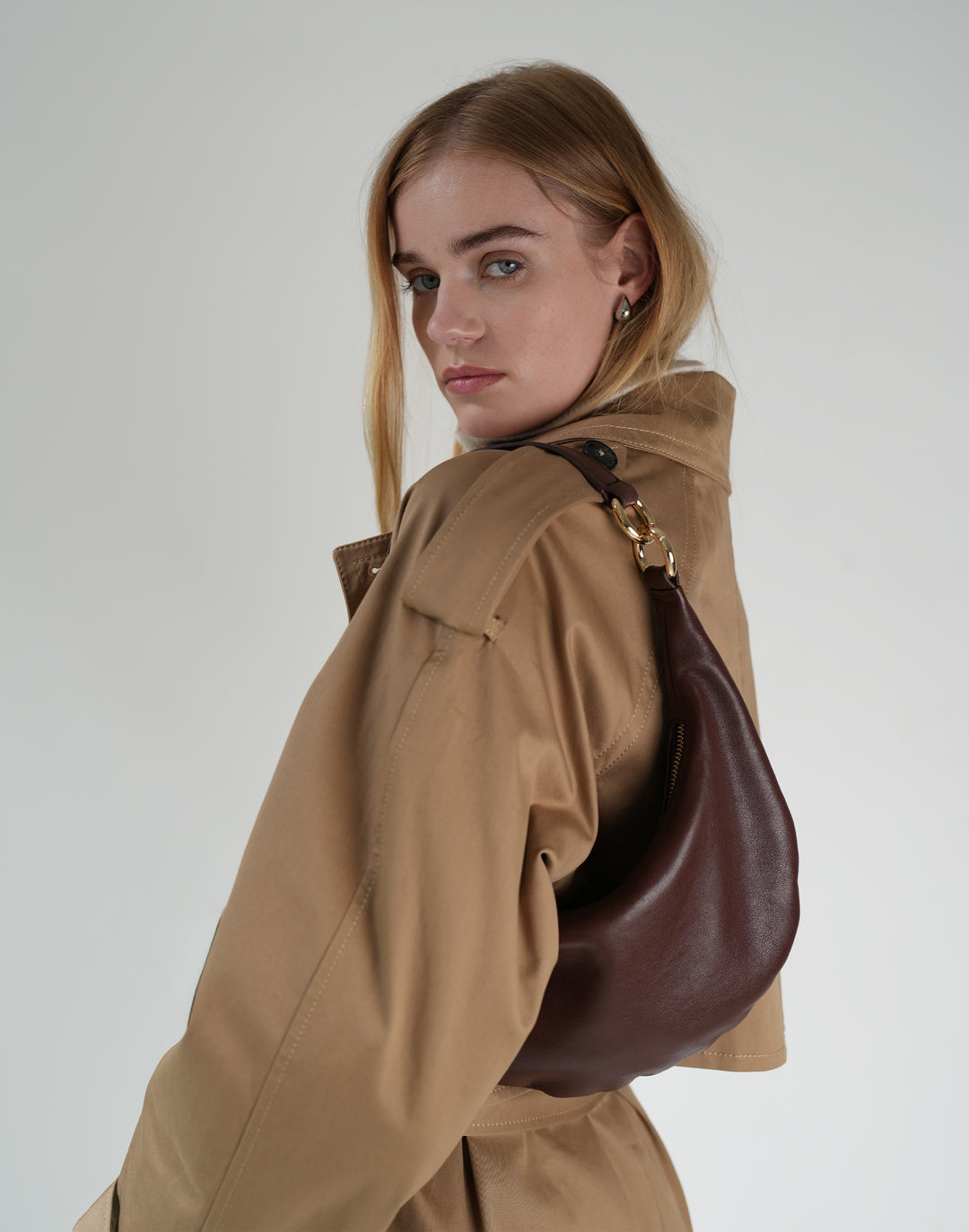a close up photo of a woman in a trench coat wearing a burgundy crescent-shaped shoulder bag with gold ring hardware #color_bordeaux-vintage
