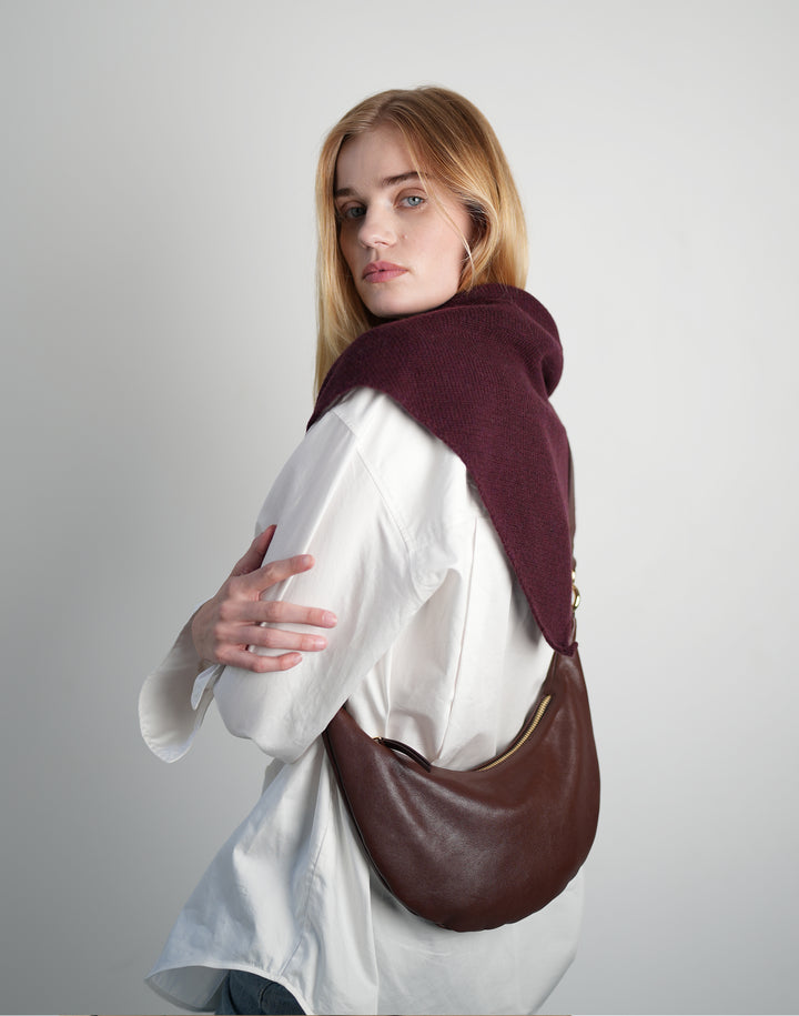 a cropped photo of a woman in a white oversized shirt with burgundy shawl wearing a burgundy crossbody crescent bag, standing against a grey background #color_bordeaux-vintage