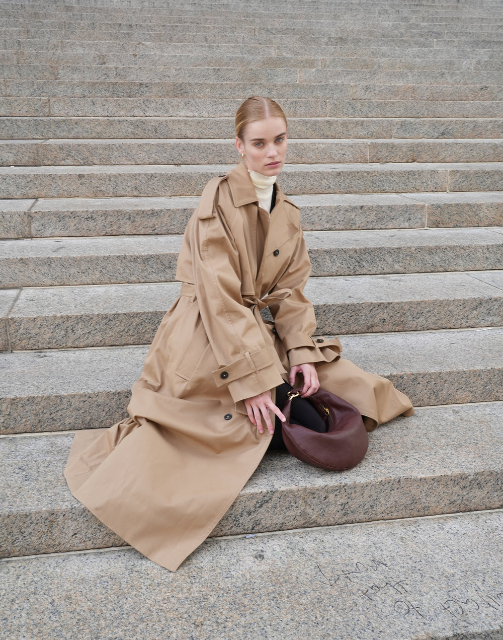 a model sits on stairs, wearing a khaki trench coat, cream turtleneck and holding a bordeaux vintage micro ring bag by HYER GOODS #color_bordeaux-vintage