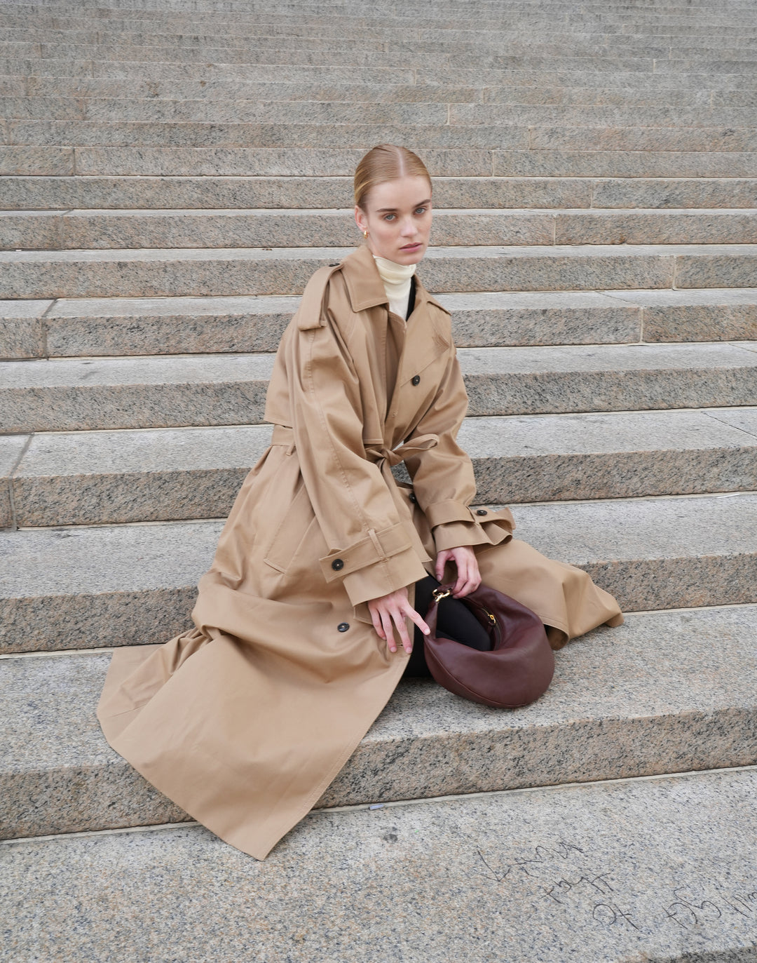 a model sits on stairs, wearing a khaki trench coat, cream turtleneck and holding a bordeaux vintage micro ring bag by HYER GOODS #color_bordeaux-vintage