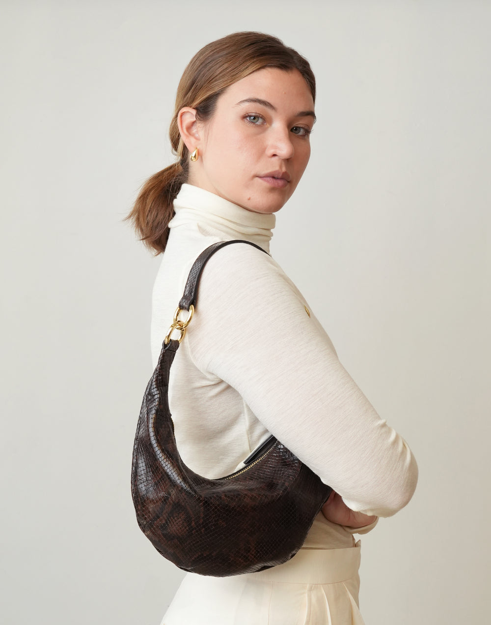 a woman wearing a cream turtleneck and cream pants wears a HYER GOODS Mini crescent micro ring shoulder bag brown python #color_brown-python-embossed