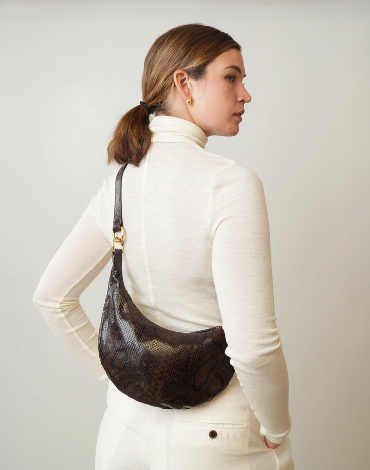 a woman wearing a cream turtleneck and cream pants wears a HYER GOODS micro ring crossbody crescent bag brown python #color_brown-python-embossed