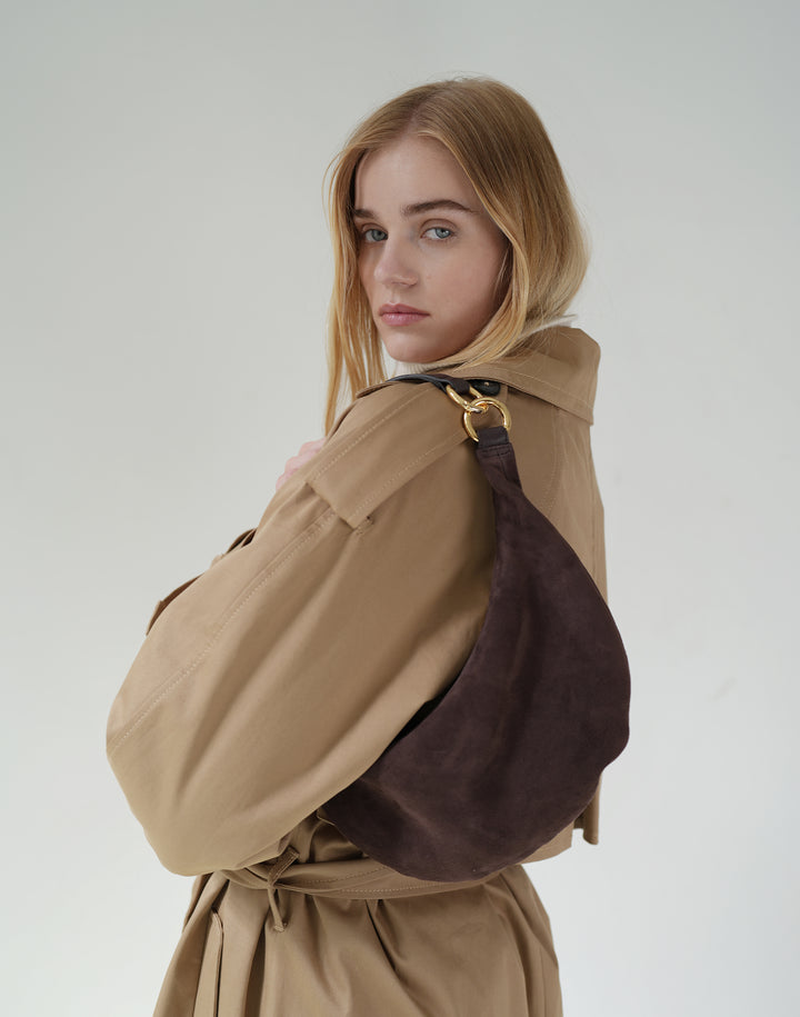 a woman in a trench coat and cream turtleneck wears a brown suede shoulder bag with gold hardware against a grey background #color_chocolate-suede