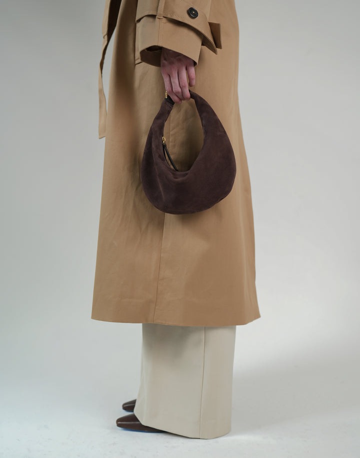 a cropped photo of the bottom half of a person wearing a trench coat, cream pants, pointy tow dark brown shoes and carrying a brown suede crescent bag in their hand #color_chocolate-suede