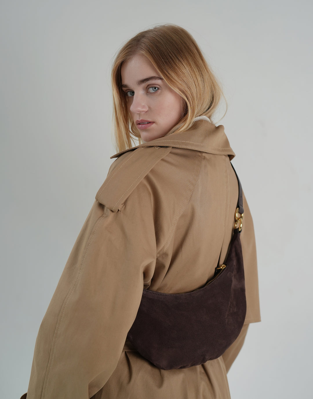 back view of a woman wearing a khaki trench, a dark brown suede crossbody crescent bag hugging her torso #color_chocolate-suede