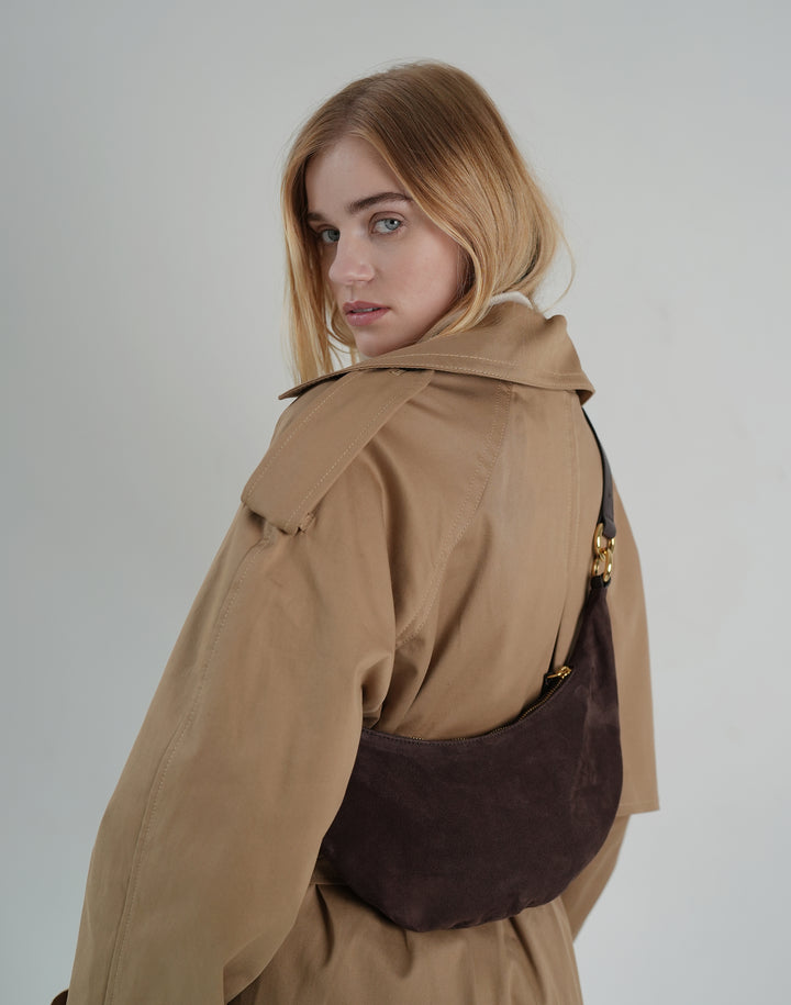 back view of a woman wearing a khaki trench, a dark brown suede crossbody crescent bag hugging her torso #color_chocolate-suede