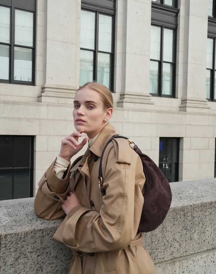 a woman in a trench coat and cream turtleneck wears a brown suede shoulder bag with gold hardware, leaning against a stone wall with a large building behind her #color_chocolate-suede