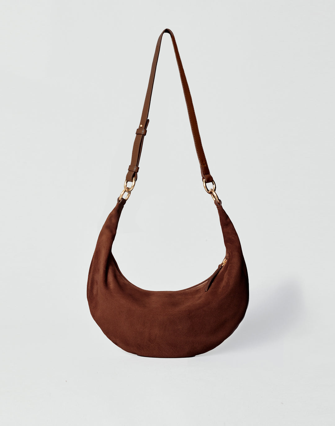 A dark brown HYER GOODS Piccola Ring Bag crescent-shaped shoulder bag made of suede, featuring a single adjustable shoulder strap with gold-tone hardware, set against a light gray background #color_chocolate-suede