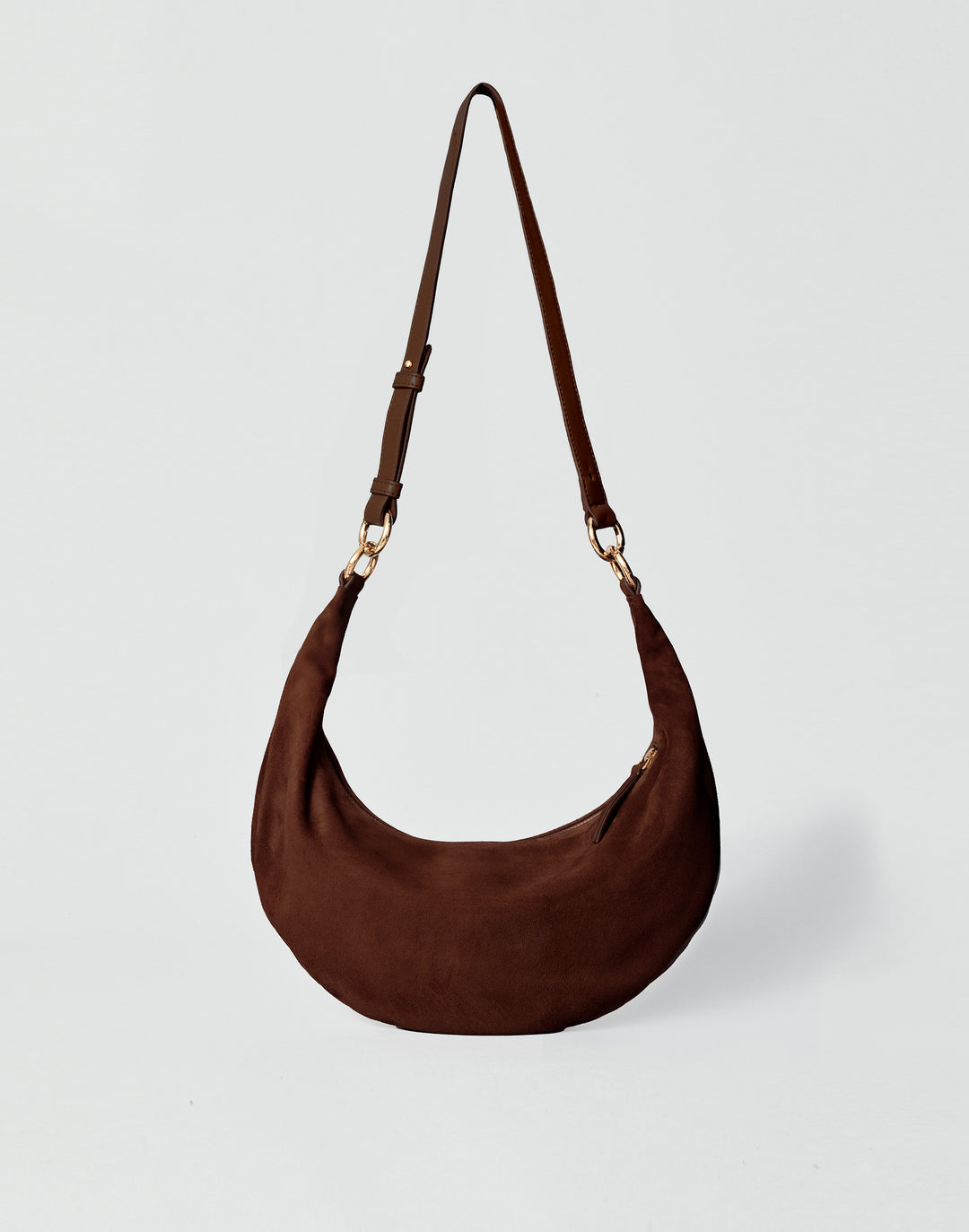 A dark brown HYER GOODS Piccola Ring Bag crescent-shaped shoulder bag made of suede, featuring a single adjustable shoulder strap with gold-tone hardware, set against a light gray background #color_chocolate-suede