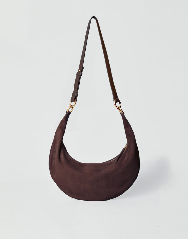 A dark brown HYER GOODS Piccola Ring Bag crescent-shaped shoulder bag made of suede, featuring a single adjustable shoulder strap with gold-tone hardware, set against a light gray background #color_chocolate-suede