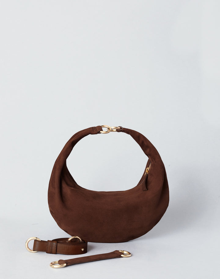 A dark brown suede crescent-shaped shoulder bag with gold-tone hardware, shown on a light gray studio background with its matching detachable strap placed in front.#color_chocolate-suede