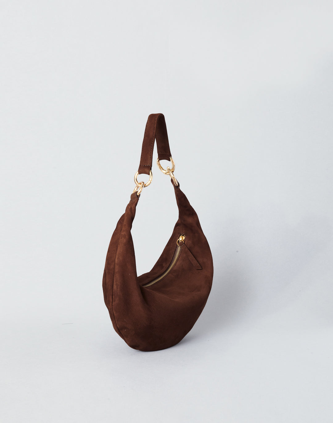 A dark brown suede crescent shoulder bag with gold-tone hardware, shown on a light gray studio background.#color_chocolate-suede