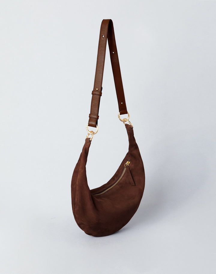 A dark brown crescent-shaped shoulder bag made of suede or faux suede, featuring a matching adjustable shoulder strap with gold-tone hardware, set against a light gray studio background.#color_chocolate-suede