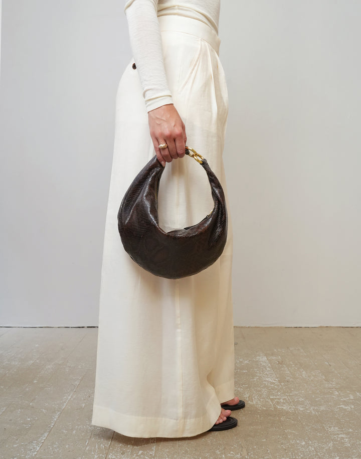 A person wearing a cream long-sleeve top and wide-leg cream trousers holds a HYER GOODS dark brown crescent-shaped dark brown python embossed leather handbag with gold hardware against a plain light gray wall and concrete floor. #color_brown-python-embossed
