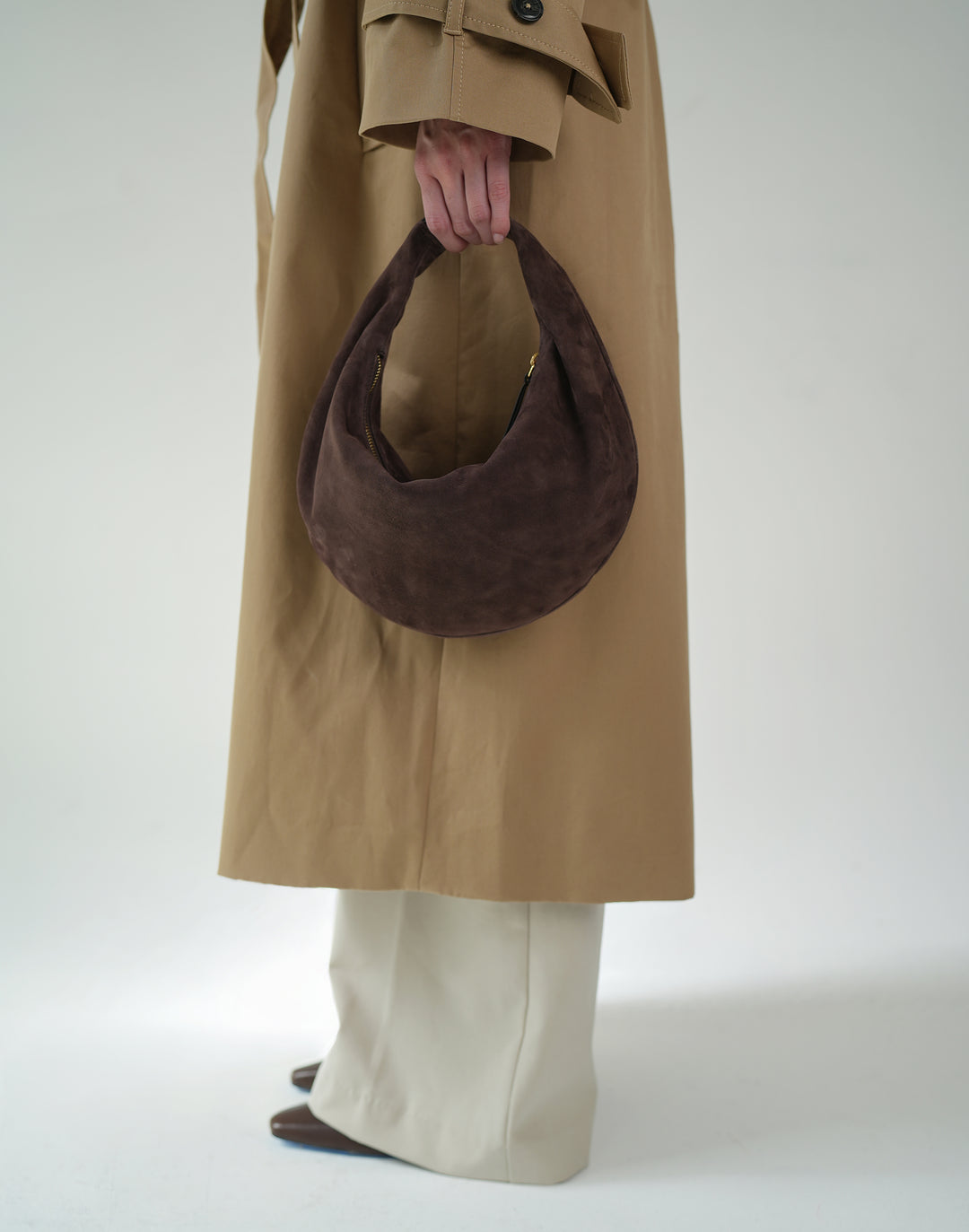 a cropped photo of a woman in cream pants, a khaki trench and a brown suede crescent bag hanging from her fingertips #color_chocolate-suede