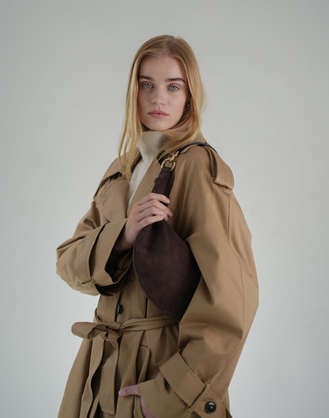 a woman wears a cream sweater turtleneck and a belted, oversized khaki trench coat with a brown suede crescent shoulder bag #color_chocolate-suede