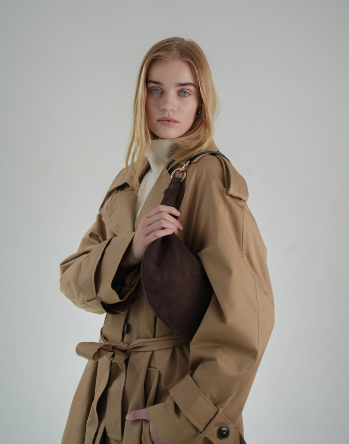 a woman wears a cream sweater turtleneck and a belted, oversized khaki trench coat with a brown suede crescent shoulder bag #color_chocolate-suede