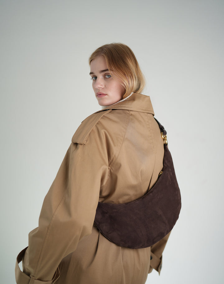 back view of a woman glancing over her shoulder at the camera, wearing an oversized khaki trench and a brown suede crossbody crescent bag across her back. #color_chocolate-suede