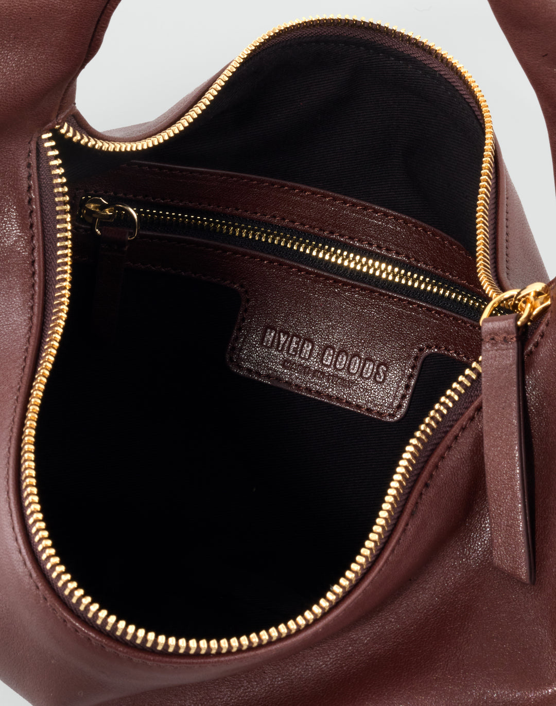 Close-up of the interior of a brown leather handbag, showing a black lined compartment with gold-tone zipper, a brown leather interior label reading HYPER GOODS, and gold zipper teeth around the opening. #color_bordeaux-vintage #color_bordeaux-vintage