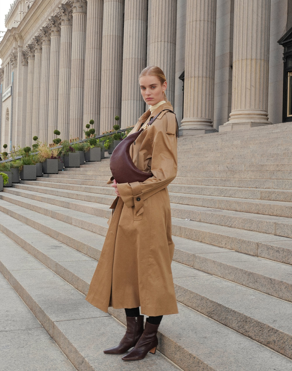 a woman in a tan trench and brown boots stands on stairs carrying a bordeaux hyer goods ring bag as a shoulder bag #color_bordeaux-vintage