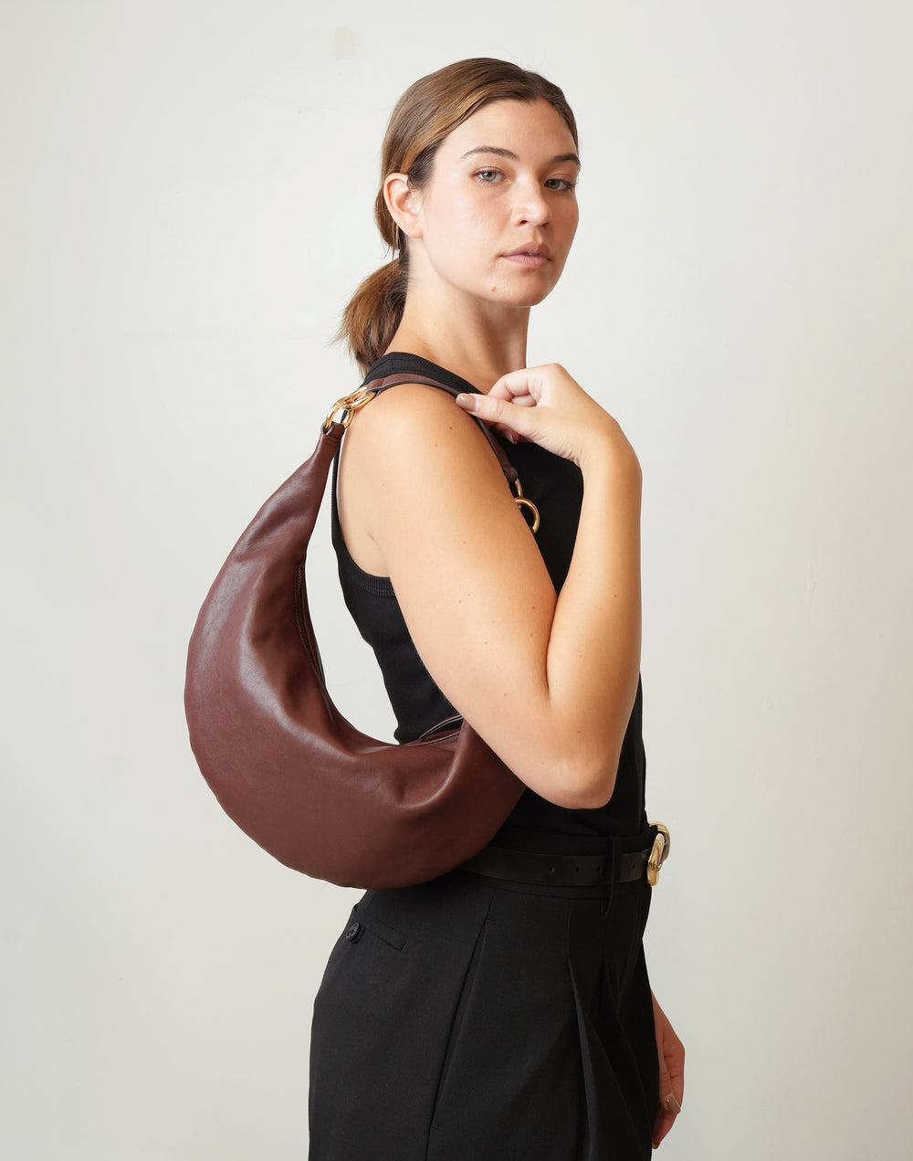 A woman wearing a black sleeveless top and black trousers carries a HYER GOODS Ring Bag large crescent-shaped red-brown leather shoulder bag with gold-tone hardware against a plain off-white background. #color_bordeaux-vintage