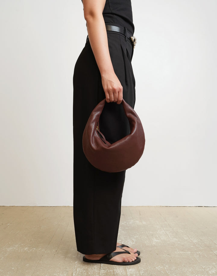 A person wearing black wide-leg trousers, a black top, and black sandals holds a HYER GOODS Rign Bag, a red-dark brown crescent-shaped leather handbag against a plain off-white wall and light floor. #color_bordeaux-vintage