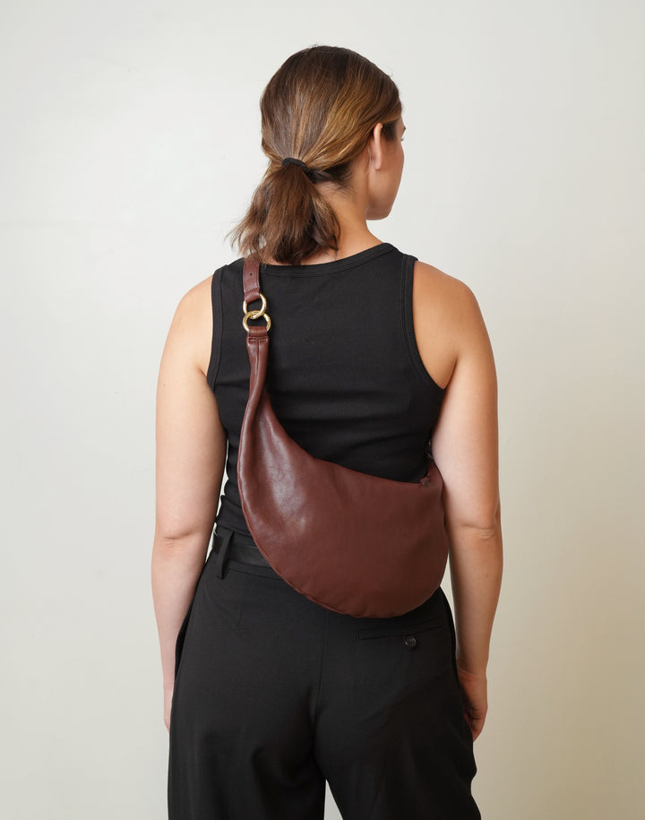 A woman viewed from behind wearing a HYER GOODS red-brown crescent-shaped leather shoulder bag with a gold-tone ring clasp over a black sleeveless top and black pants, against a plain off-white background. #color_bordeaux-vintage