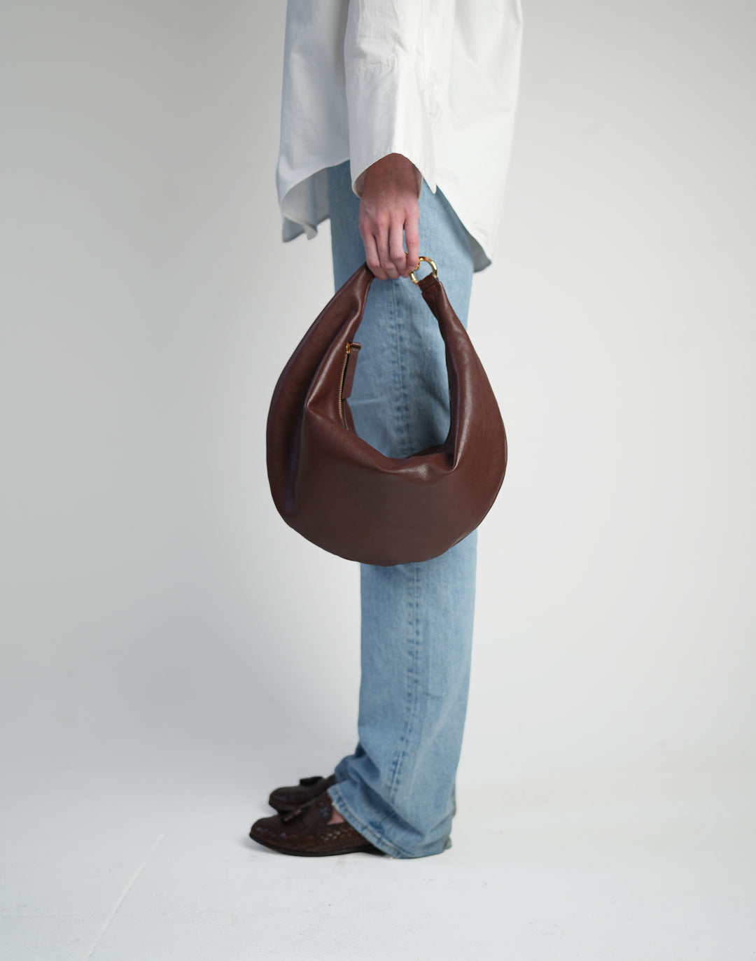 a profile of a person, cropped from the waist down, wearing a loose white button down shirt, light jeans, brown loafers and holding a bordeaux hyer goods ring bag in their fingertips #color_bordeaux-vintage