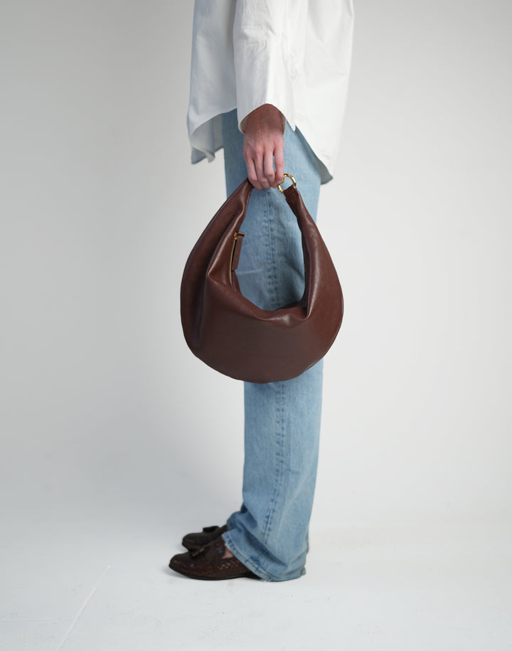 a profile of a person, cropped from the waist down, wearing a loose white button down shirt, light jeans, brown loafers and holding a bordeaux hyer goods ring bag in their fingertips #color_bordeaux-vintage