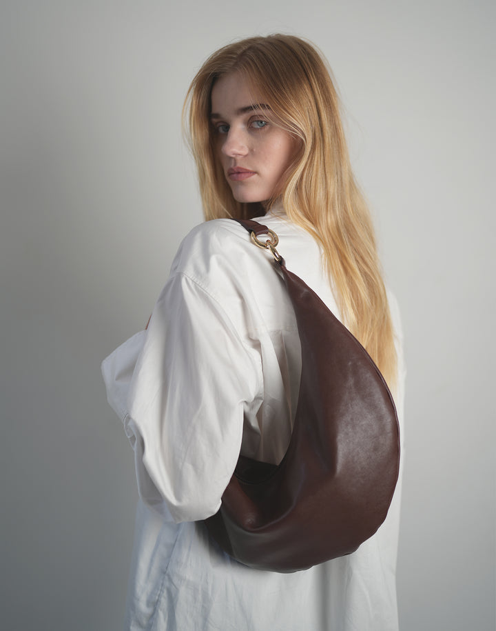 a blond woman wearing an oversized white shirt stands with her back to the camera, wearing a burgundy shoulder bag with gold ring hardware #color_bordeaux-vintage
