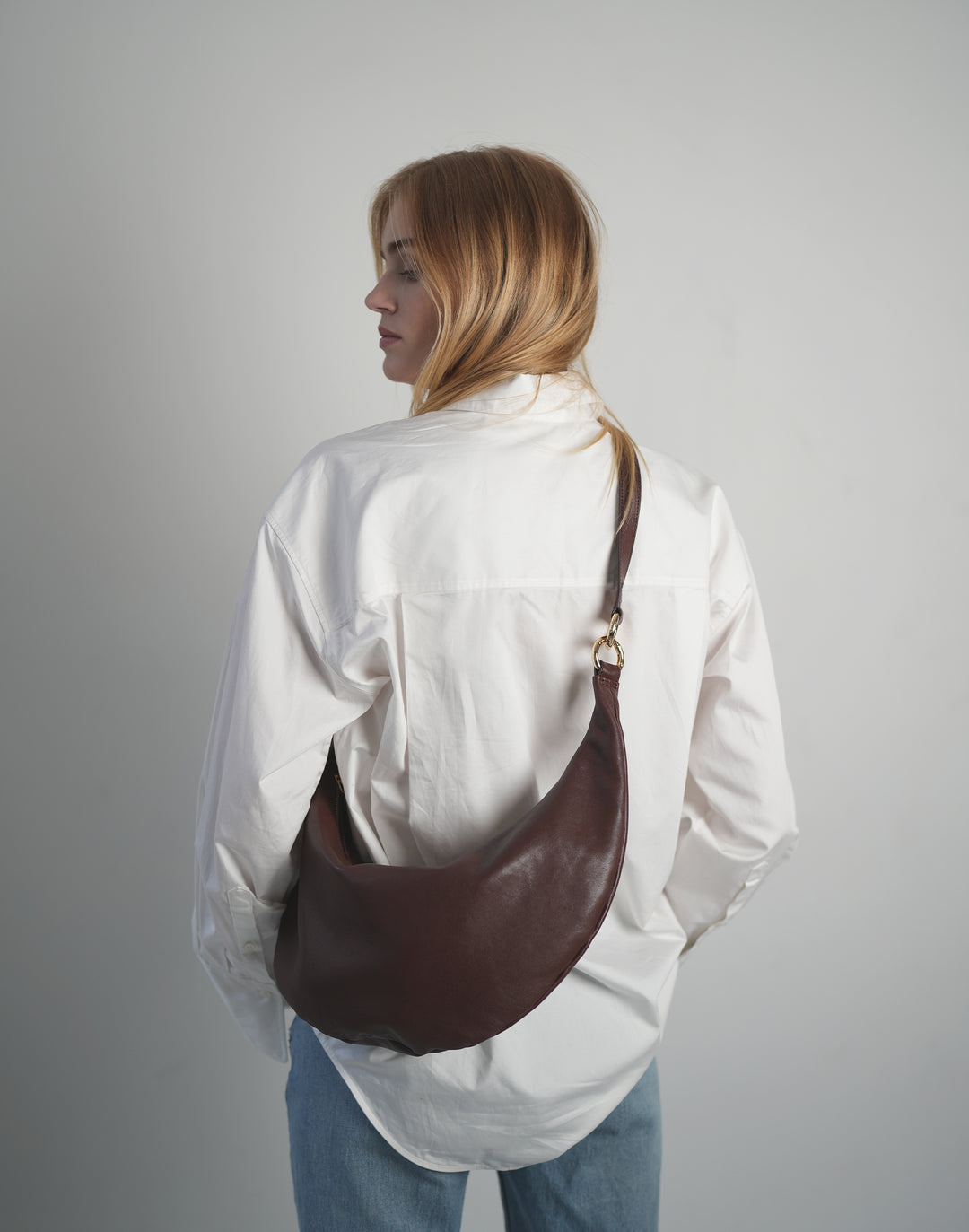 a woman in an oversized white shirt and jeans stands with her back to the camera, with a burgundy crossbody crescent bag draped across her back #color_bordeaux-vintage