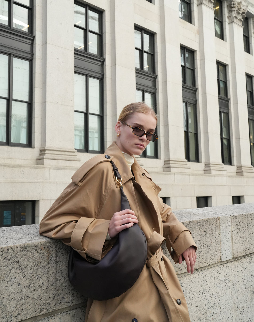 a woman in a trench coat and cream turtleneck leans agains a city wall, holding a dark brown leather crescent-shaped shoulder bag with gold hardware on her shoulder #color_chocolate