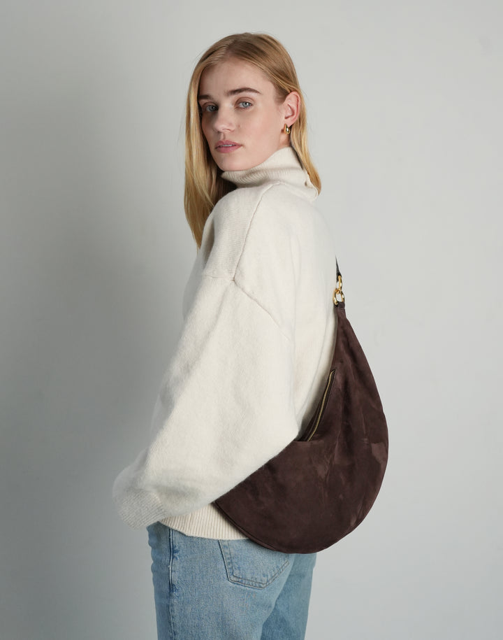 profile shot of a woman in an oversized cream sweater, light blue jeans, and a slouchy dark brown suede crossbody crescent bag with gold hardware #color_chocolate-suede
