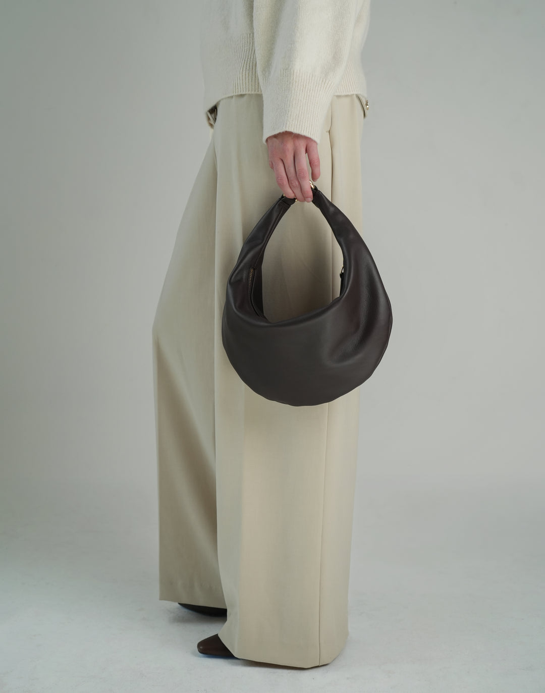 a cropped shot of a woman's light khaki pants and brown pointy boots, she holds a brown leather crescent bag in her hand #color_chocolate