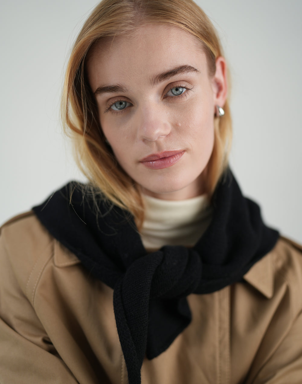 a cropped photo of a woman with blond hair and blue eyes wearing a black neck scarf, cream turtleneck and khaki trench coat against a grey background #color_black