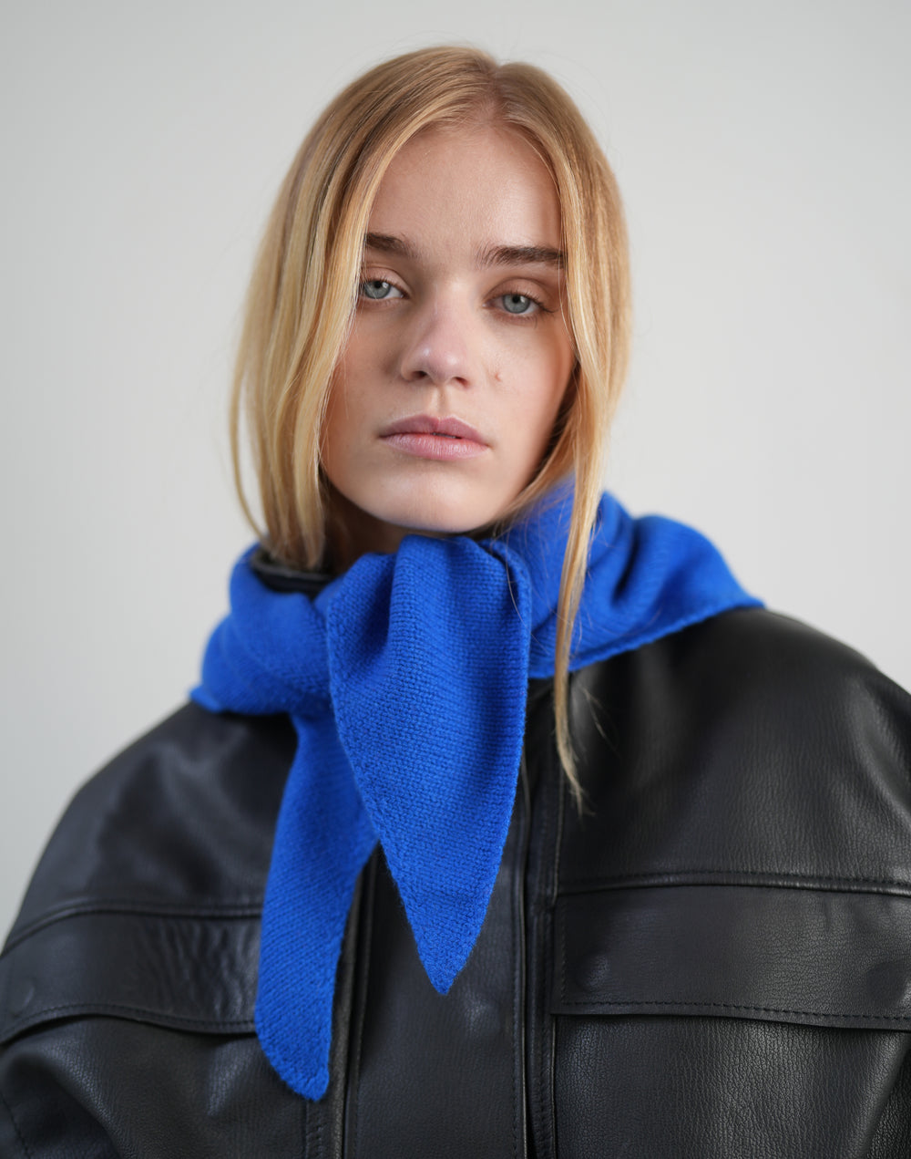 A blonde woman wearing a bright royal blue knit scarf tied around her neck, paired with a black leather jacket, against a plain light gray background. #color_cobalt