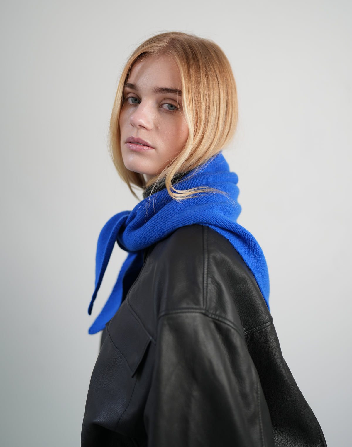 A blonde woman wearing a bright royal blue knit scarf and a black leather jacket, photographed against a neutral light gray studio background.#color_cobalt