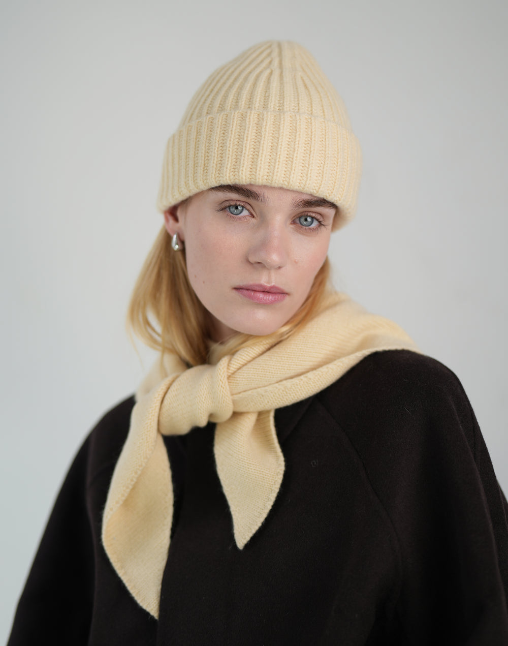 a cropped photo of a woman in a light yellow-colored ribbed beanie and a matching scarf tied around her neck with a dark brown coat against a grey background #color_butter-yellow
