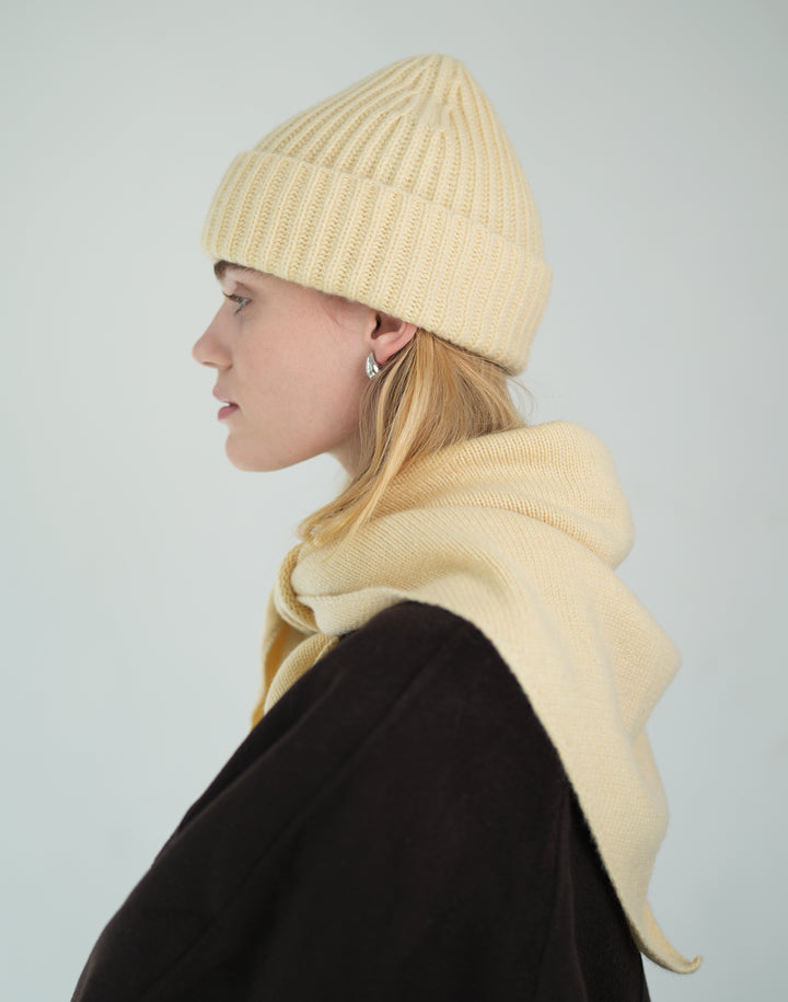 A woman shown in profile wearing a cream-colored ribbed knit beanie and a matching cream knit hooded scarf or shawl, paired with a dark brown coat, against a light gray studio background. #color_butter-yellow