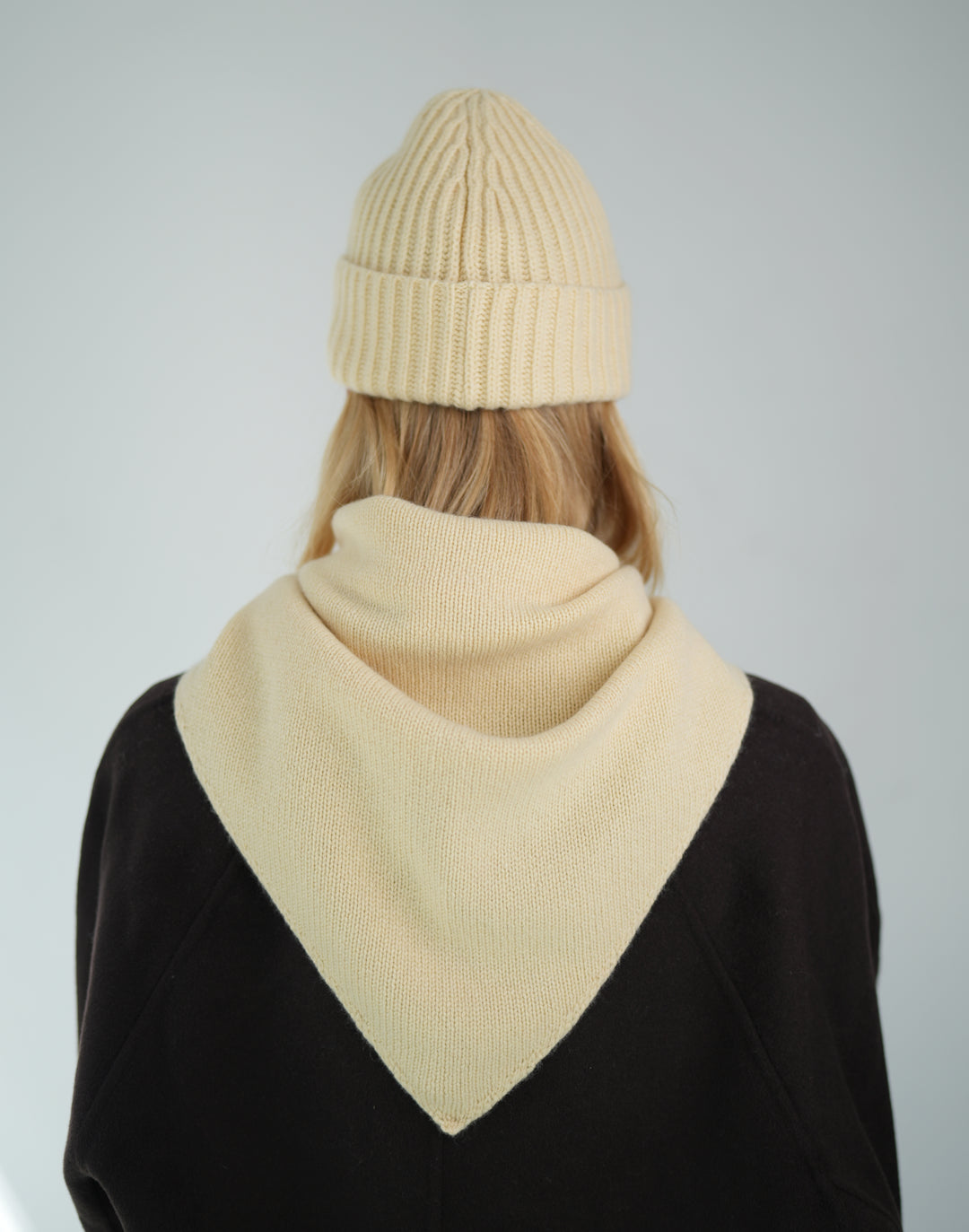 A person shown from the back wearing a cream ribbed knit beanie and a matching cream cowl neck scarf over a black top, against a light gray studio background. #color_butter-yellow