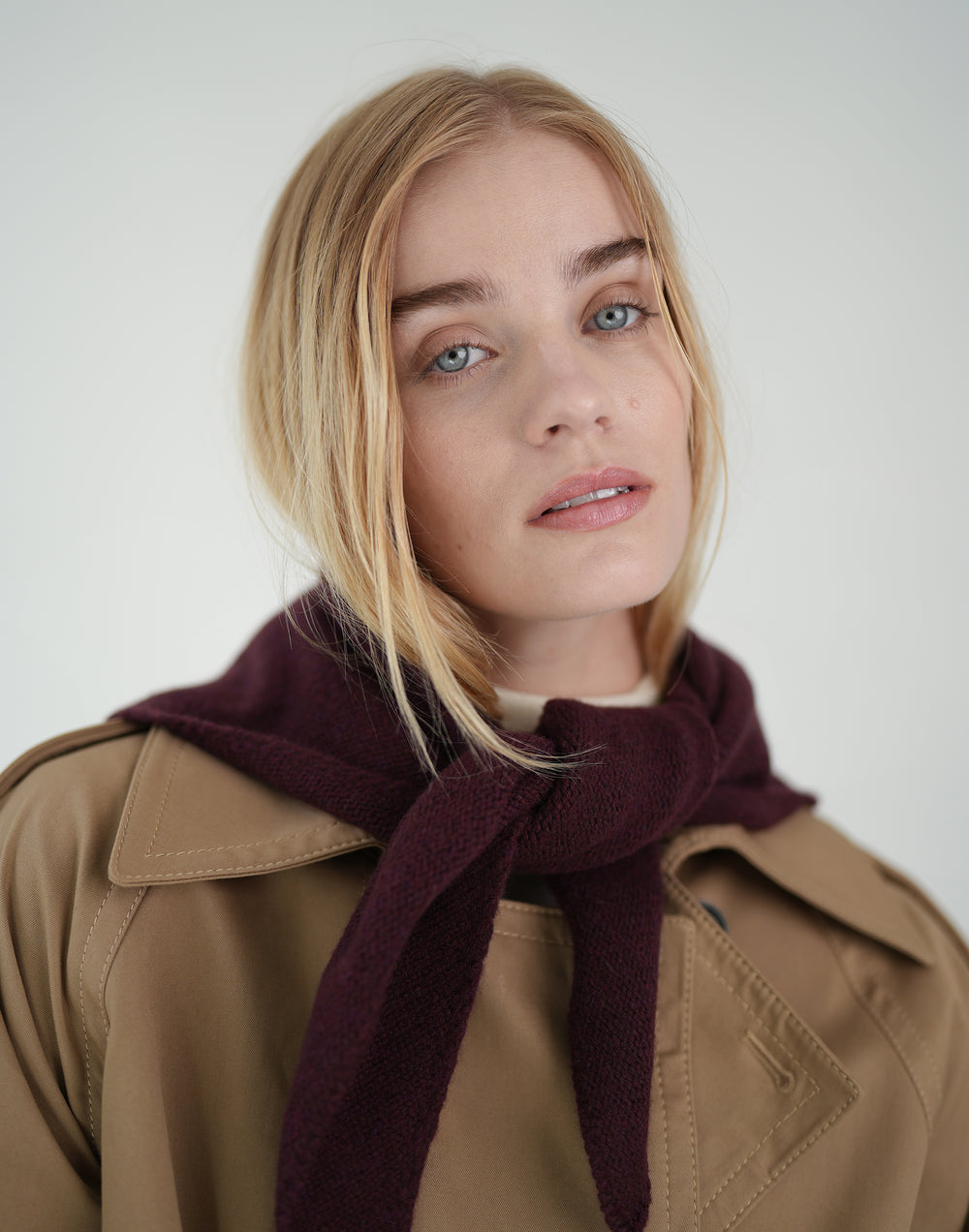 A blonde woman wearing a tan trench coat and a dark burgundy knit scarf, photographed against a plain light gray studio background. #color_bordeaux