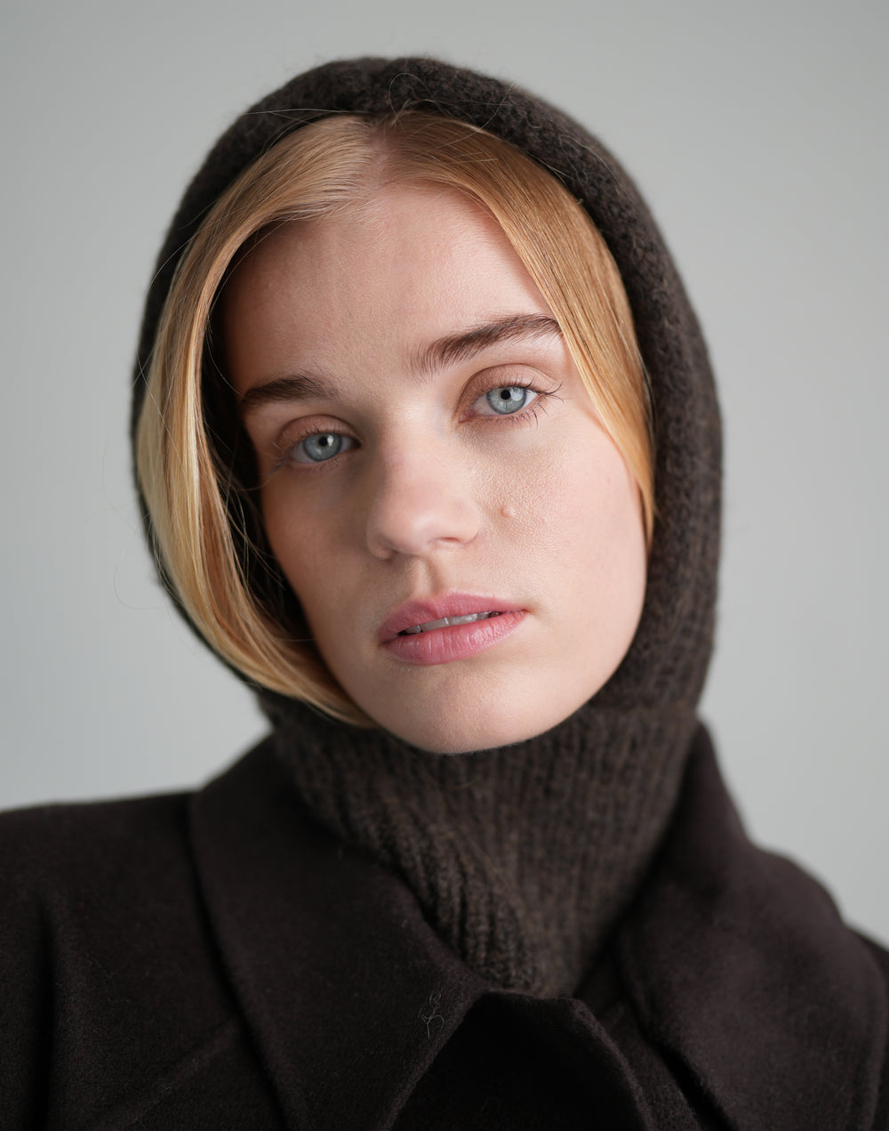 A close-up portrait of a woman wearing a dark brown knit hooded turtleneck sweater and a dark brown coat against a neutral gray background. #color_brown