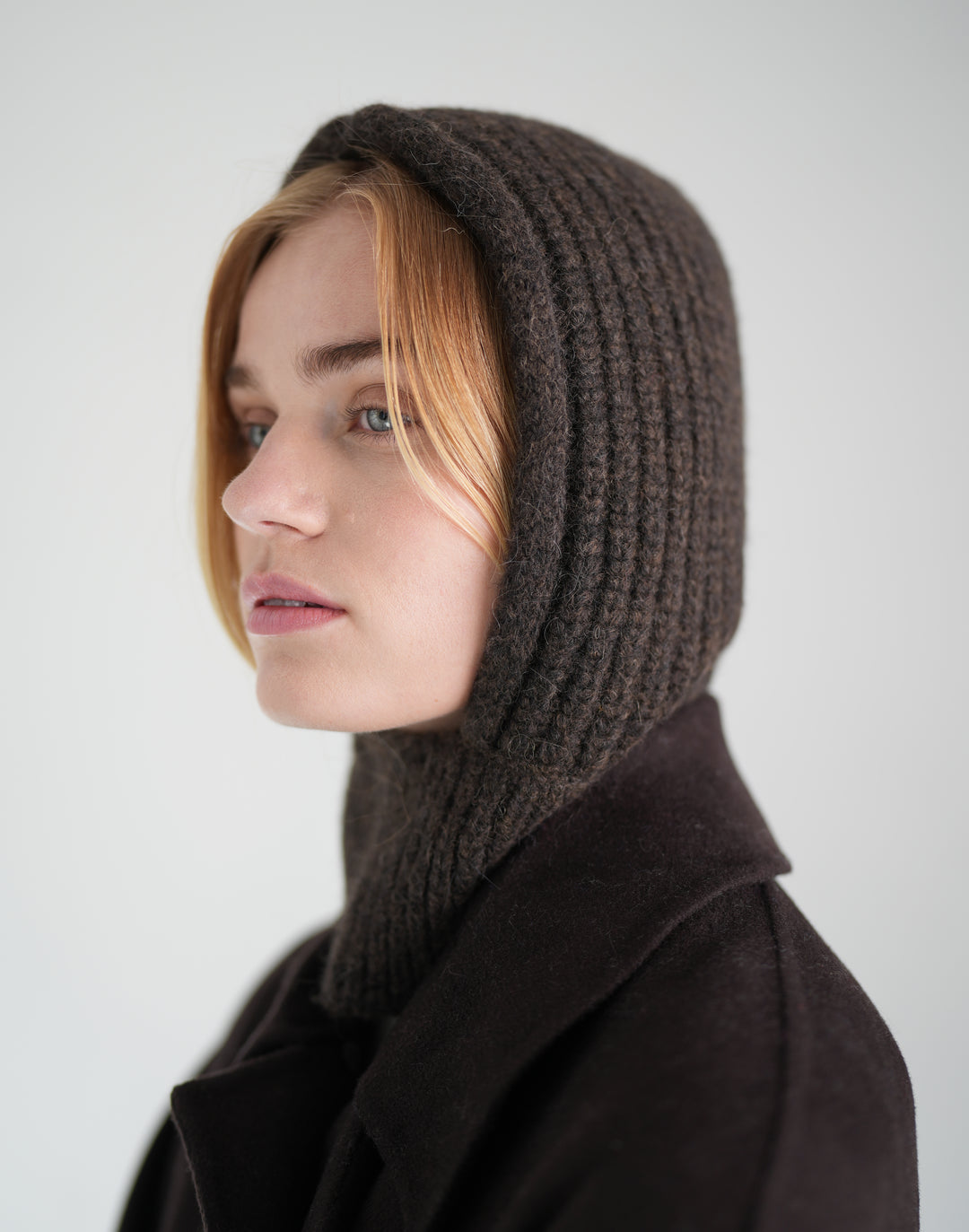 A woman in profile wearing a dark brown ribbed knit hood with an integrated neck warmer, paired with a dark coat, against a plain light gray background. #color_brown