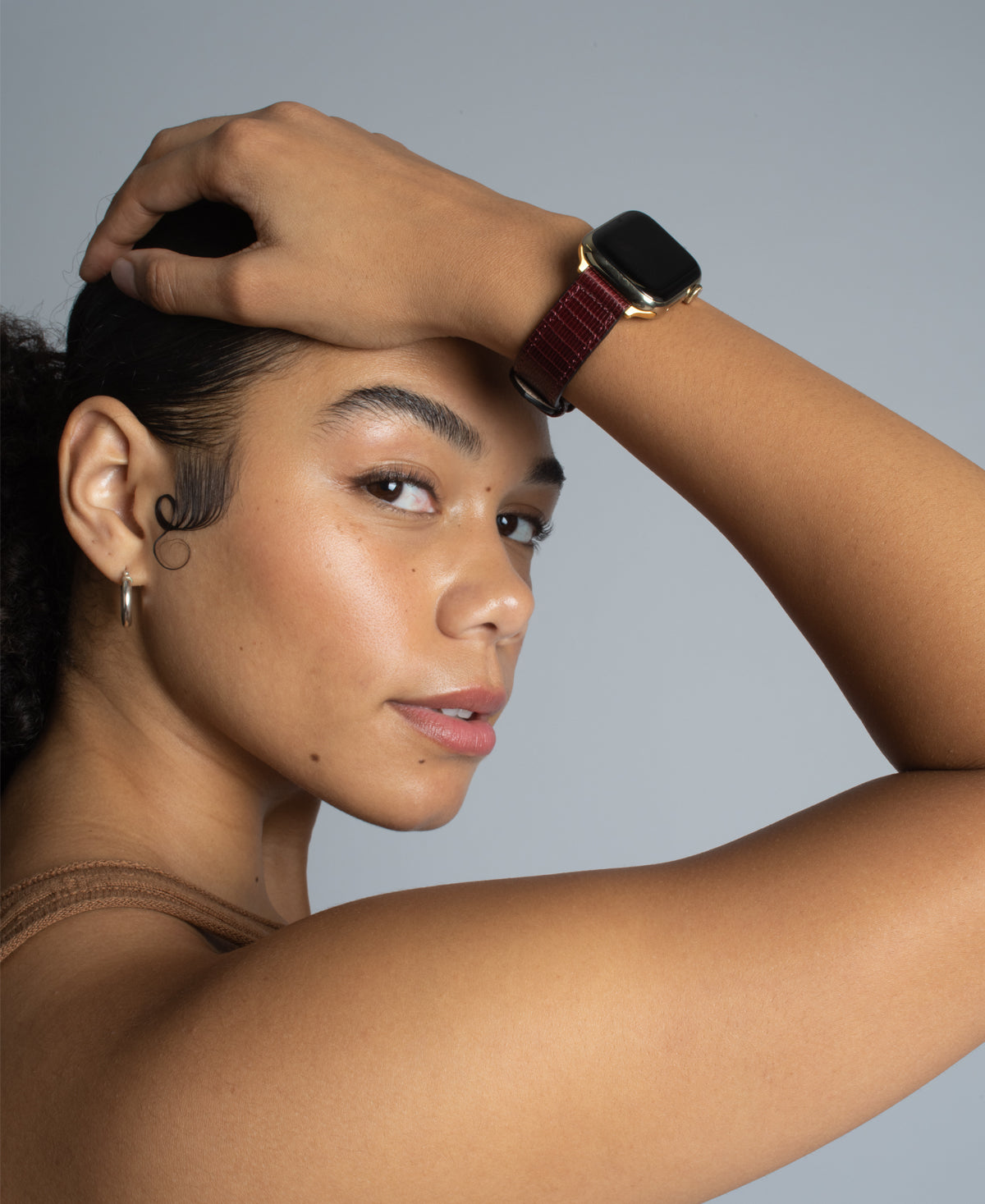 a girl wears with her hand on her head, looking sideways at the camera, wearing an apple watch with a cherry red lizard leather band 