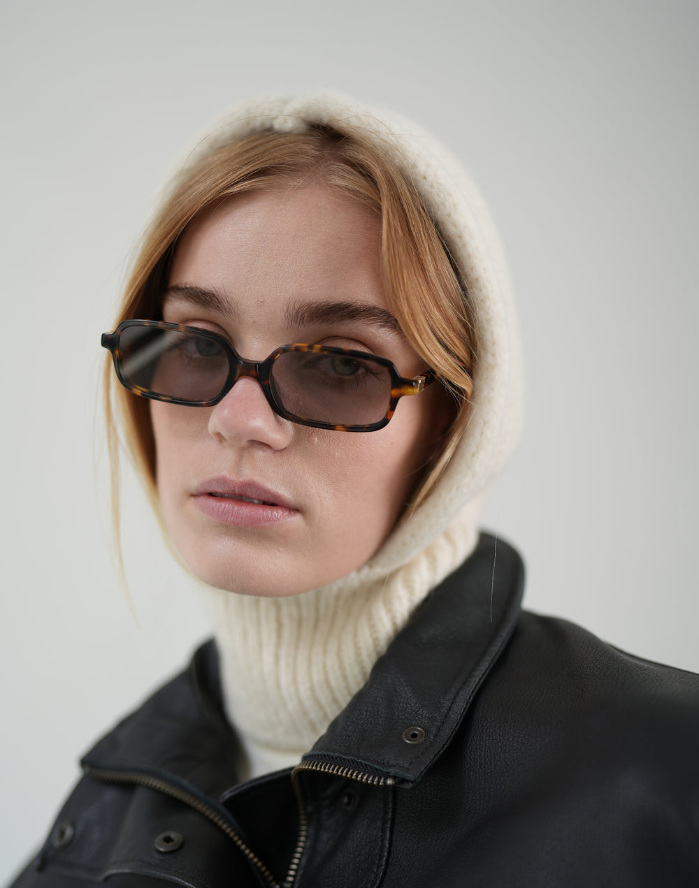 a close up image of a womans face, wearing brown sunflasses, a cream ribbed balaclava with her strawberry blond hair peeking out, and a black leather jacket #color_cream