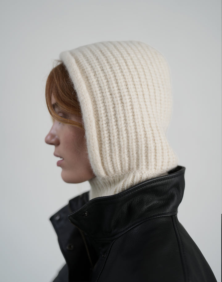 A person shown in side profile wearing a cream ribbed knit hooded balaclava over reddish-brown hair, paired with a black leather jacket, against a plain light gray background. #color_cream