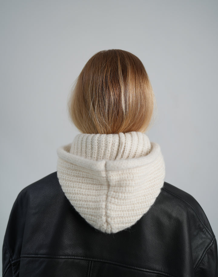 the back of a person with strawberry blond hair wearing a cream ribbed hooded balaclava down as a hood, over a black leather jacket, shot against a grey backdrop #color_cream