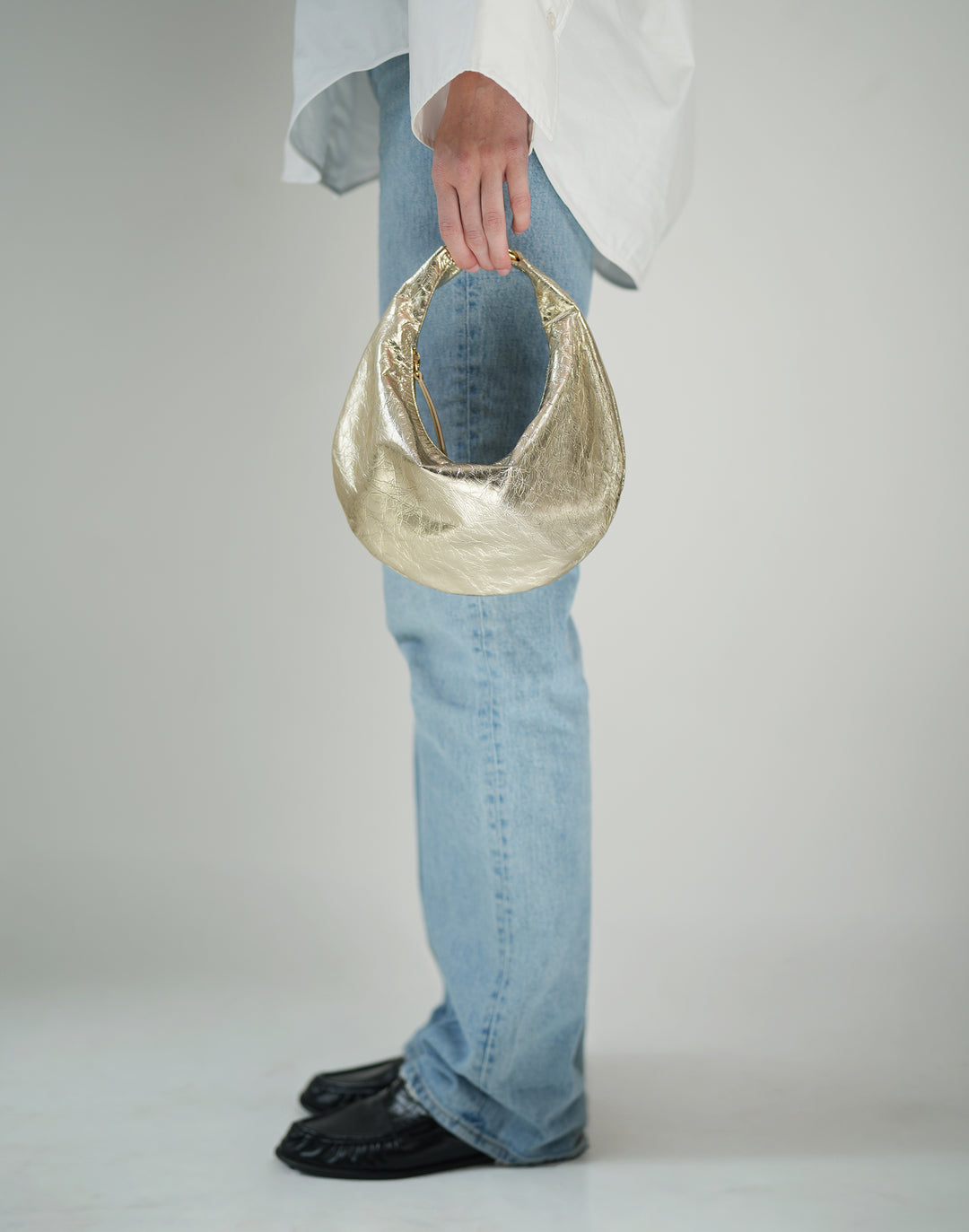 cropped profile photo of a woman's jeans, black loafers and in her hand is a gold leather crescent bag #color_gold